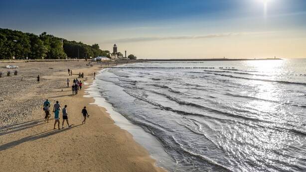 Una playa con gente caminando por la orilla, un faro al fondo y un cielo despejado.