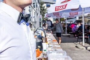 A person in a white shirt and bow tie standing in front of a buffet table with food items and a large white umbrella with 'TYSKIE' branding.