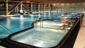 Una piscina interior con zona de hidromasaje, rodeada de sillones de descanso y una terraza de madera.