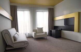 Quarto de hotel moderno com sofá, poltrona, TV de tela plana e uma janela grande com cortinas.