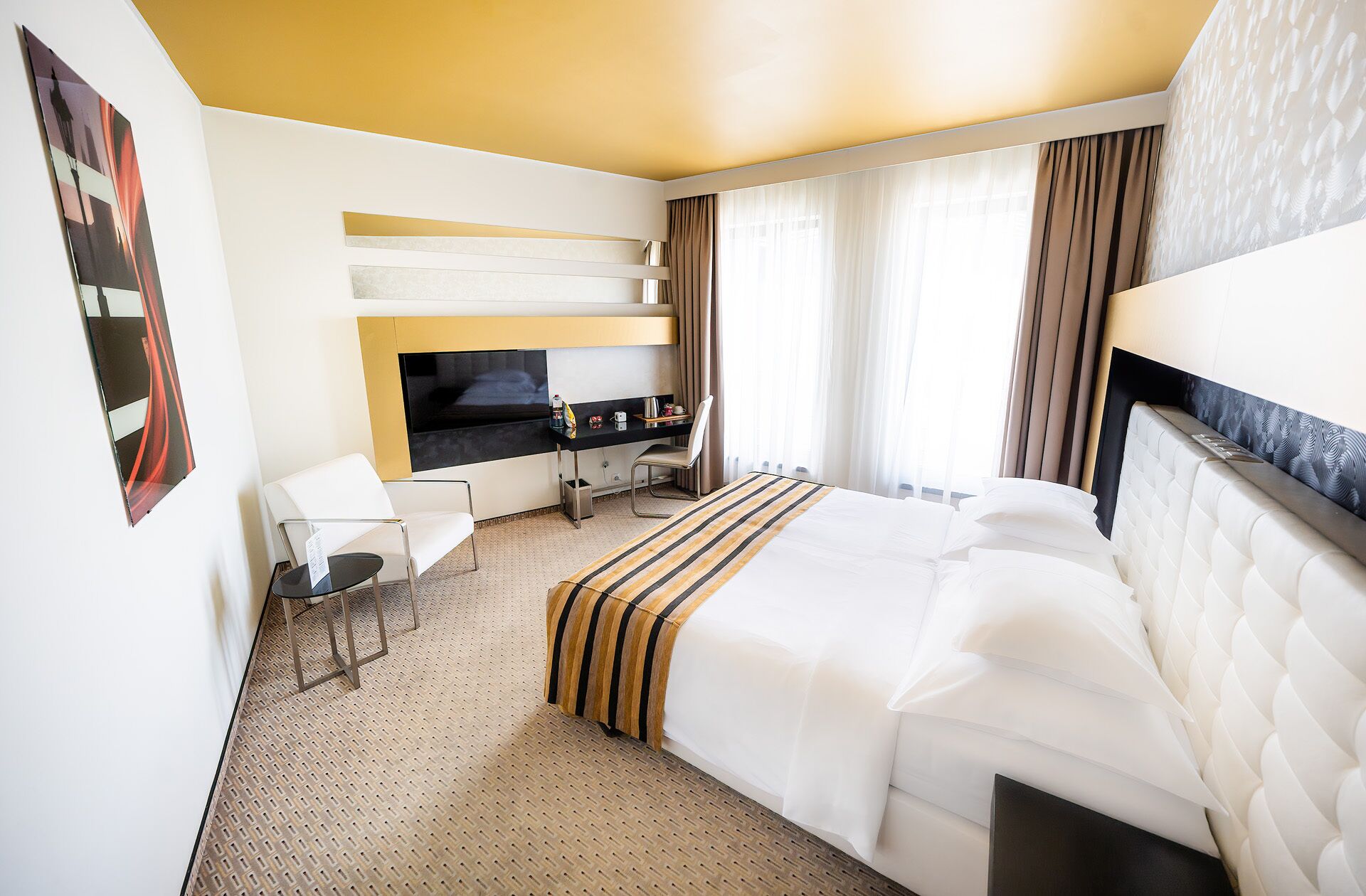 A hotel room with a bed, a desk with a chair, a TV, and a window with curtains.