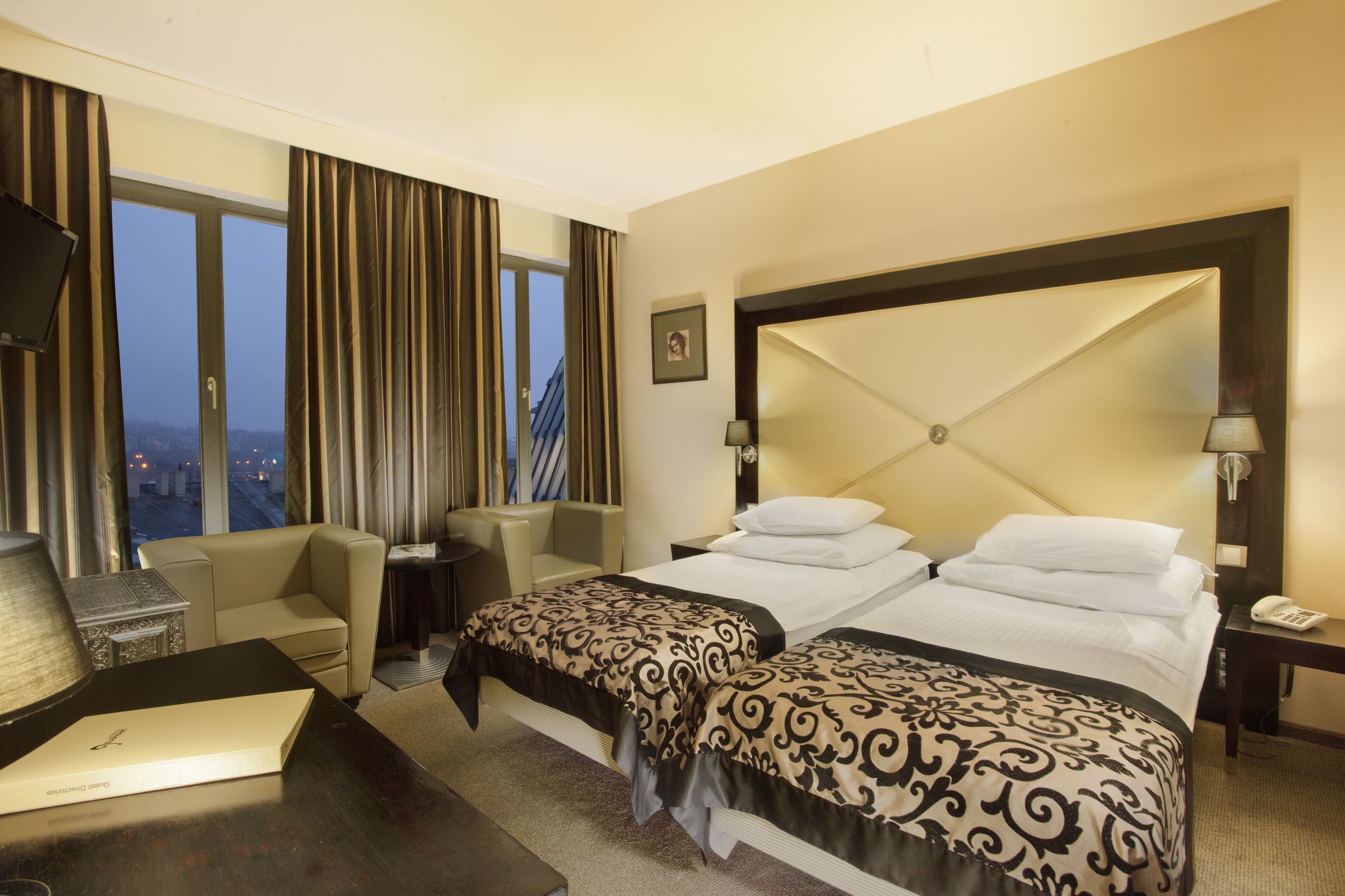 Ein Hotelzimmer mit einem großen Bett, zwei Nachttischen, einem Schreibtisch mit Lampe, einem Sessel und Blick auf die Stadt durch große Fenster.