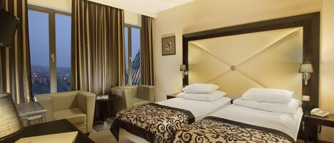 A hotel room with a large bed, two bedside tables, a desk with a lamp, a chair, and a view of the city through large windows.