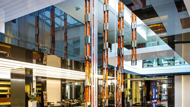 A modern lobby with a glass ceiling, a central art installation, and a seating area with a checkered floor pattern.