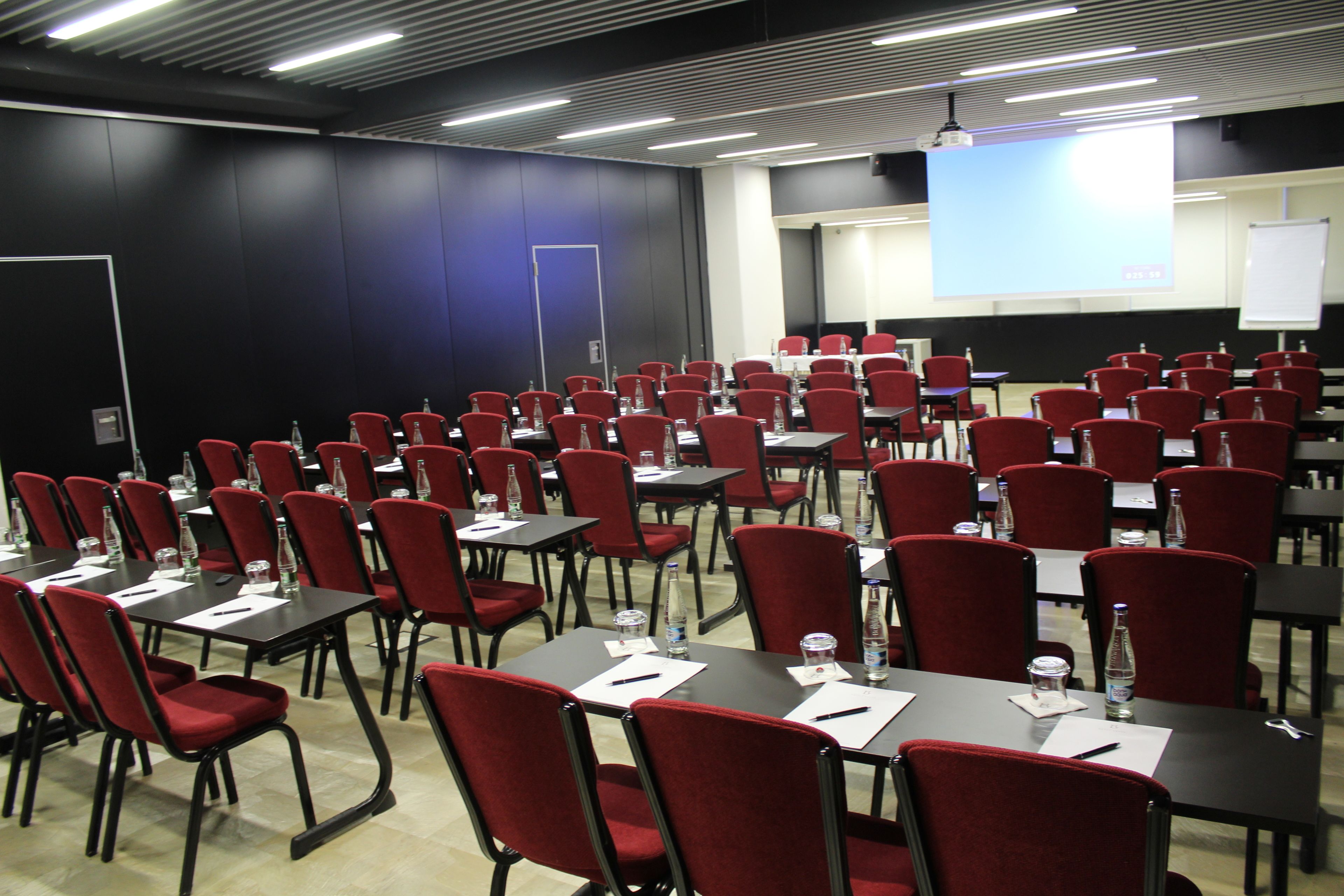 Uma sala de conferências com fileiras de cadeiras vermelhas e mesas, equipada com garrafas de água e cartões de identificação.