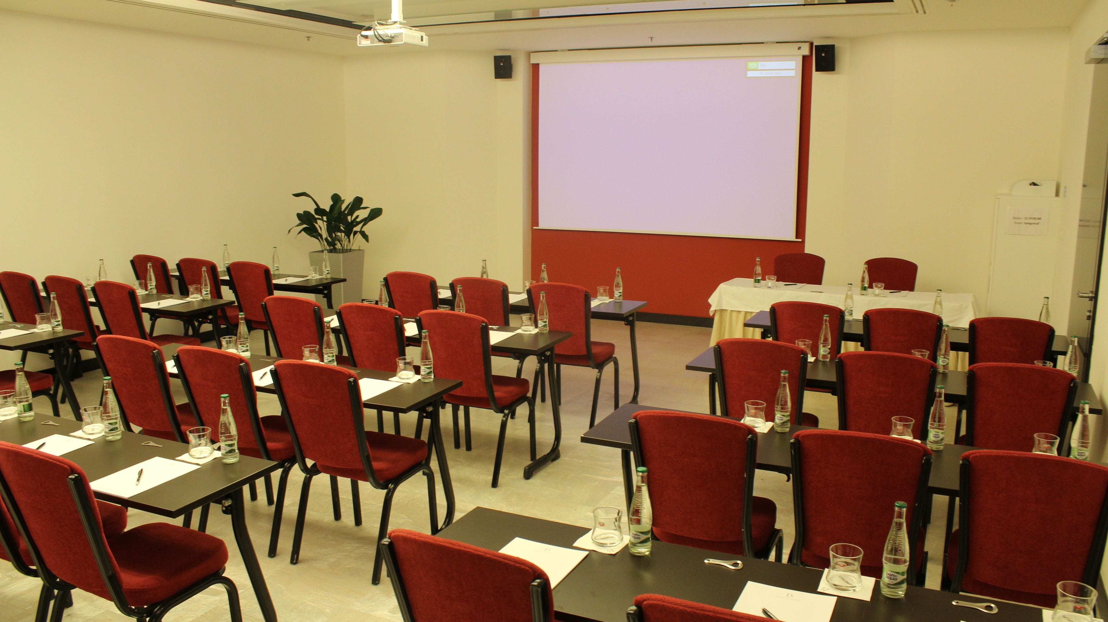 A conference room with red chairs and tables, a projector screen, and a potted plant.