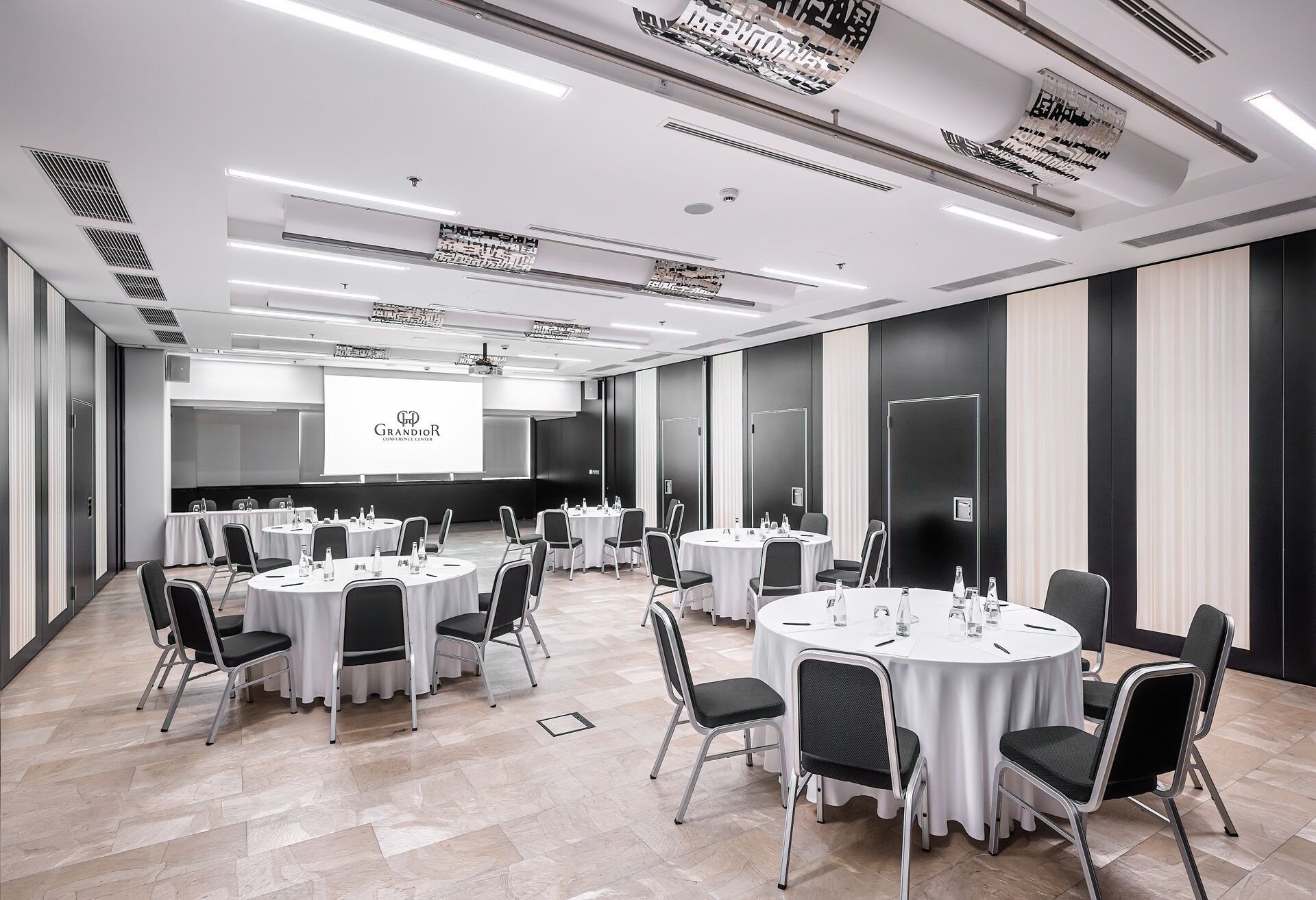 A conference room with multiple round tables, chairs, and a projection screen displaying 'Grandior'.