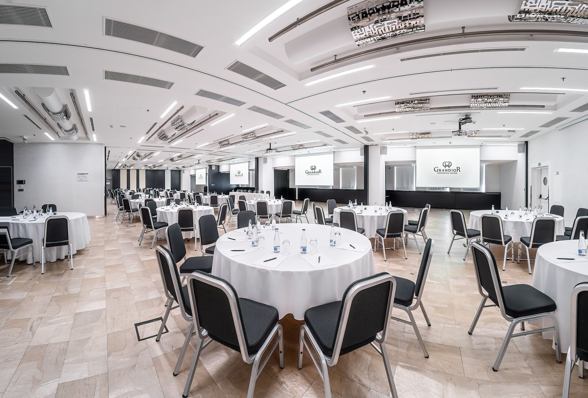 A conference room with multiple round tables, black chairs, and white tablecloths.
