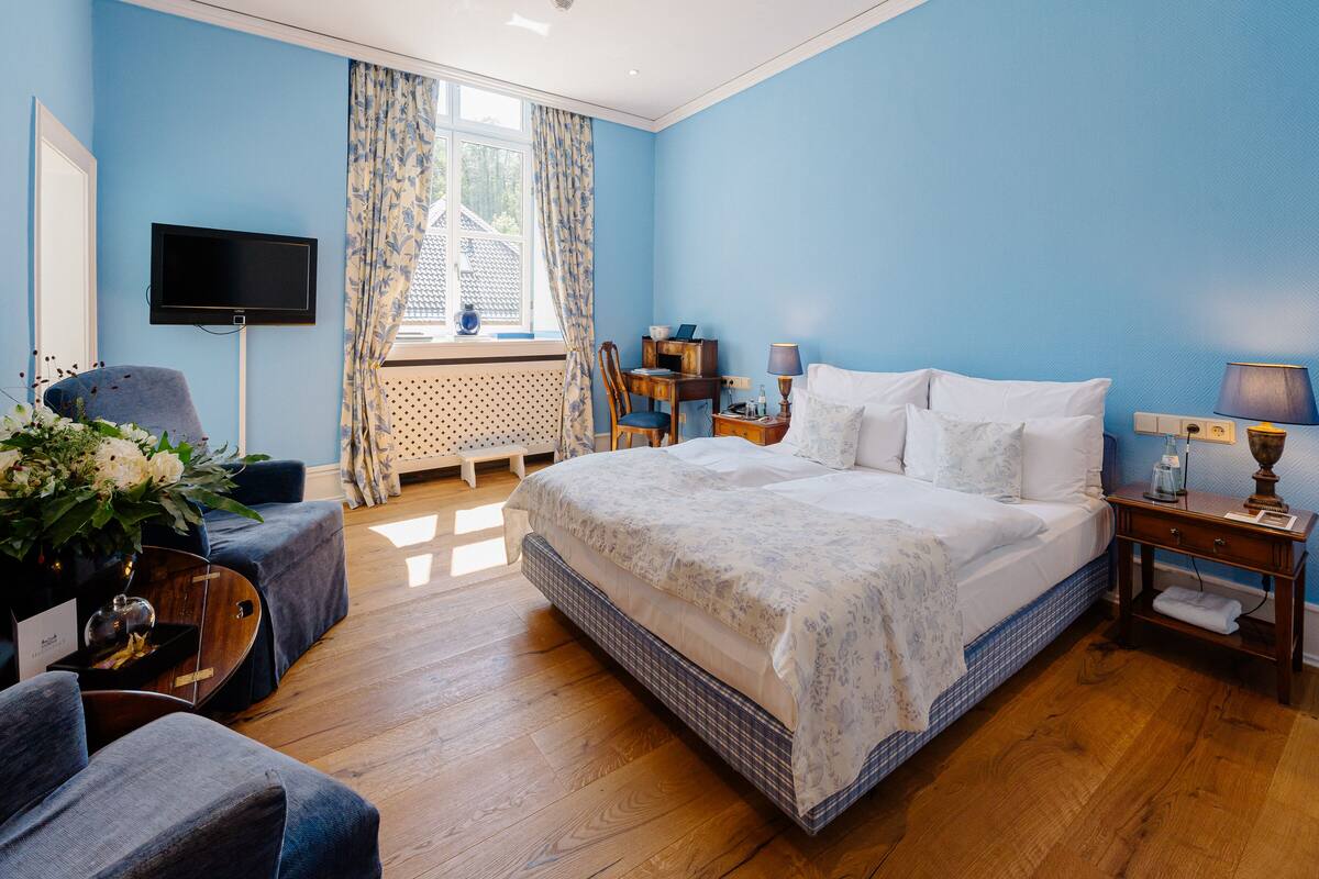 Een slaapkamer met een groot bed, een bureau, een stoel, een televisie en een raam met gordijnen.