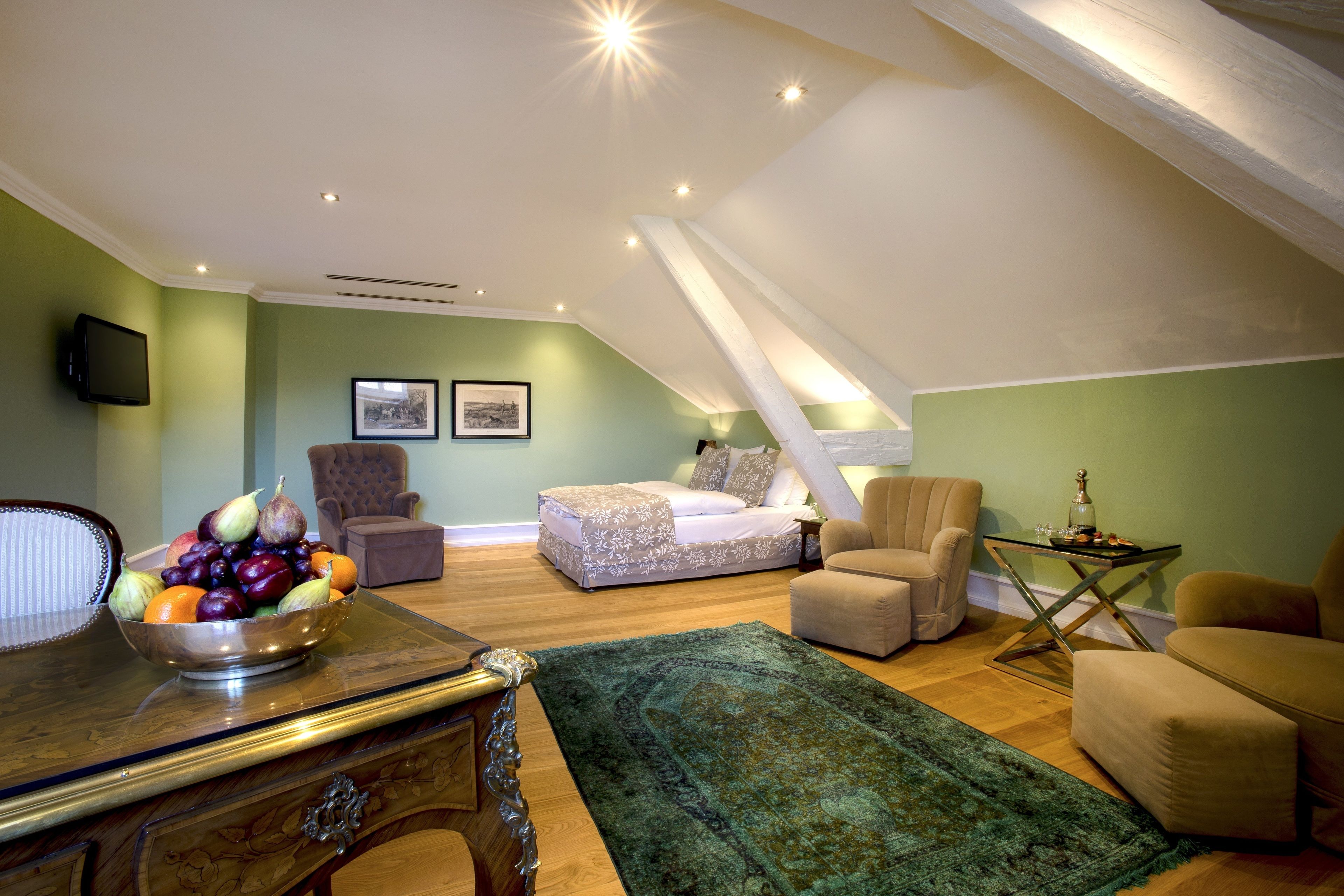 A cozy attic bedroom with a bed, two armchairs, a dining table with fruit, and a green rug.