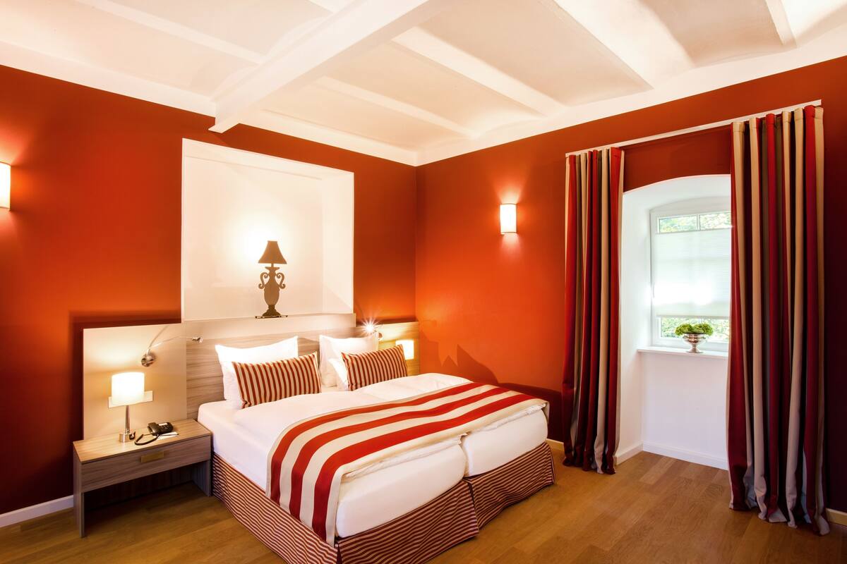 Een hotelkamer met een groot bed, een nachtkastje met een lamp en een raam met gordijnen.