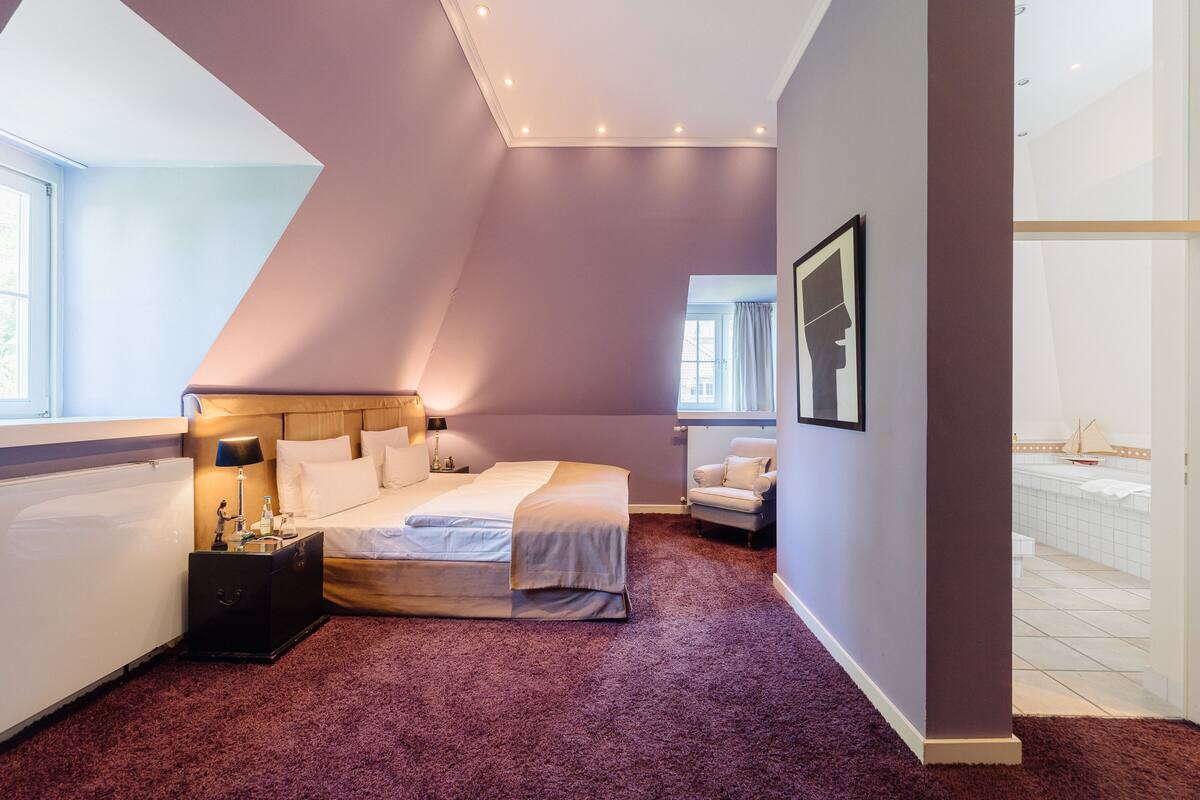 Een slaapkamer met een schuine kap, een bed met witte lakens, een nachtkastje met een lamp, en uitzicht op een badkamer met een bad.
