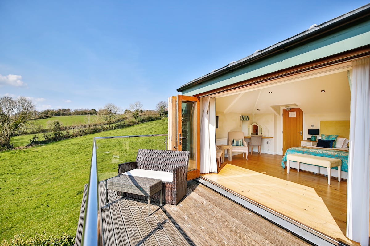 A spacious balcony with a wooden deck, a wicker sofa, and a dining table. Inside, there is a bedroom with a bed, a bedside table, and a bathroom with a shower.