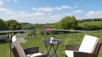 A balcony with wicker chairs and a small table, overlooking a green landscape with trees and a clear sky.