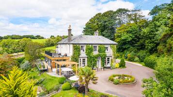 A large, historic house with a spacious patio, surrounded by lush greenery and a well-maintained garden.