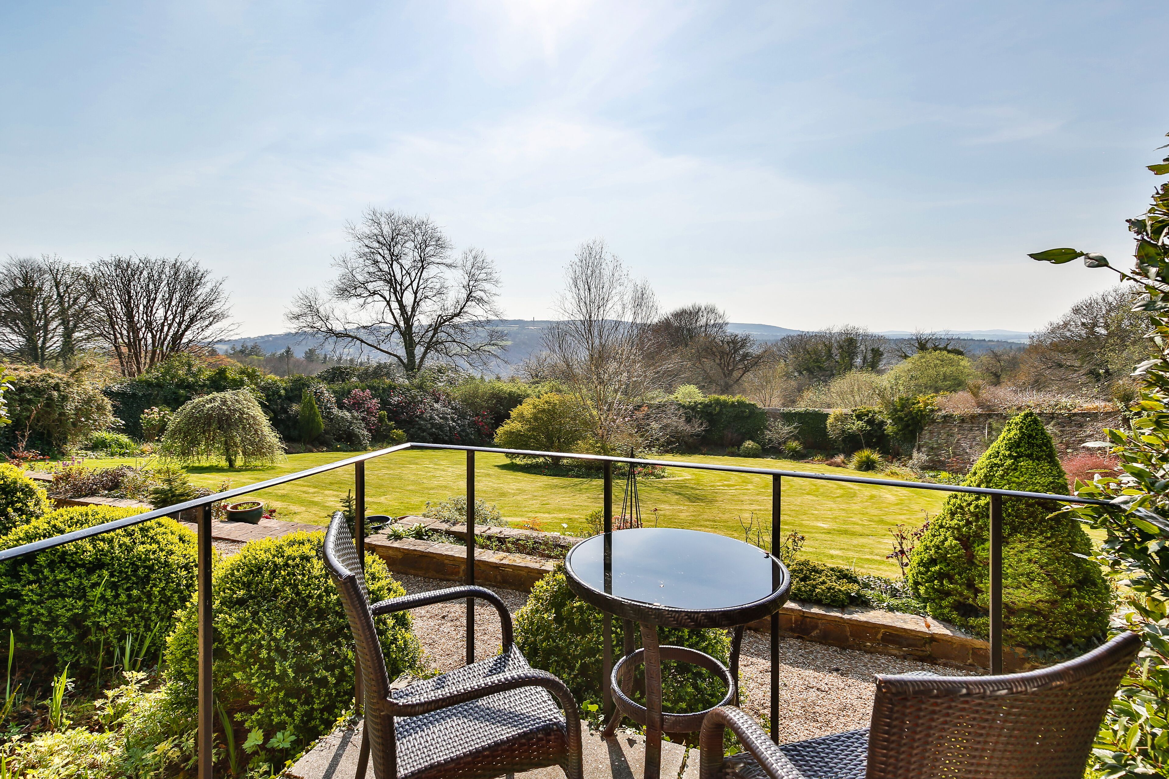 A balcony with wicker chairs and a table overlooking a well-manicured garden and distant hills.