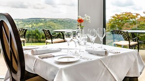 A dining table set for four with a white tablecloth, a vase with flowers, and a scenic view of hills and trees through a large window. - The Horn of Plenty (Tavistock)