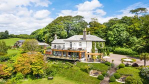 A large house with a spacious patio area, surrounded by lush greenery and a well-maintained garden. - The Horn of Plenty (Tavistock)