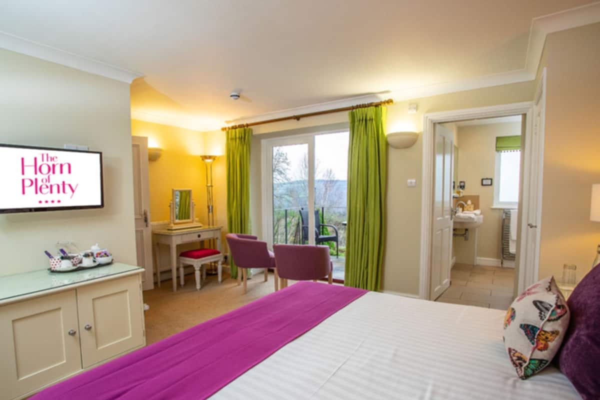 A hotel room with a bed, a desk, chairs, a bathroom, and a balcony with curtains.