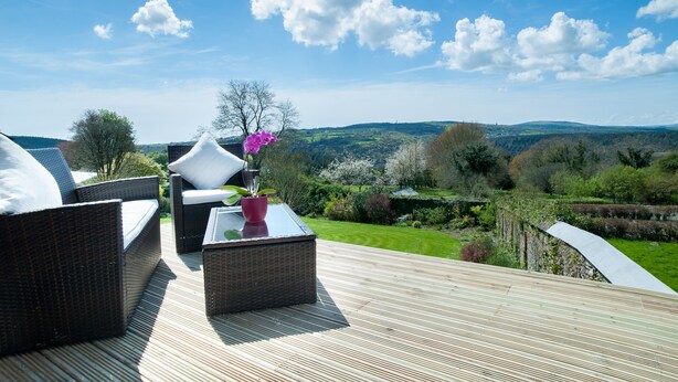 A deck with wicker furniture and a potted plant, overlooking a landscaped garden and distant hills. - The Horn of Plenty (Tavistock)