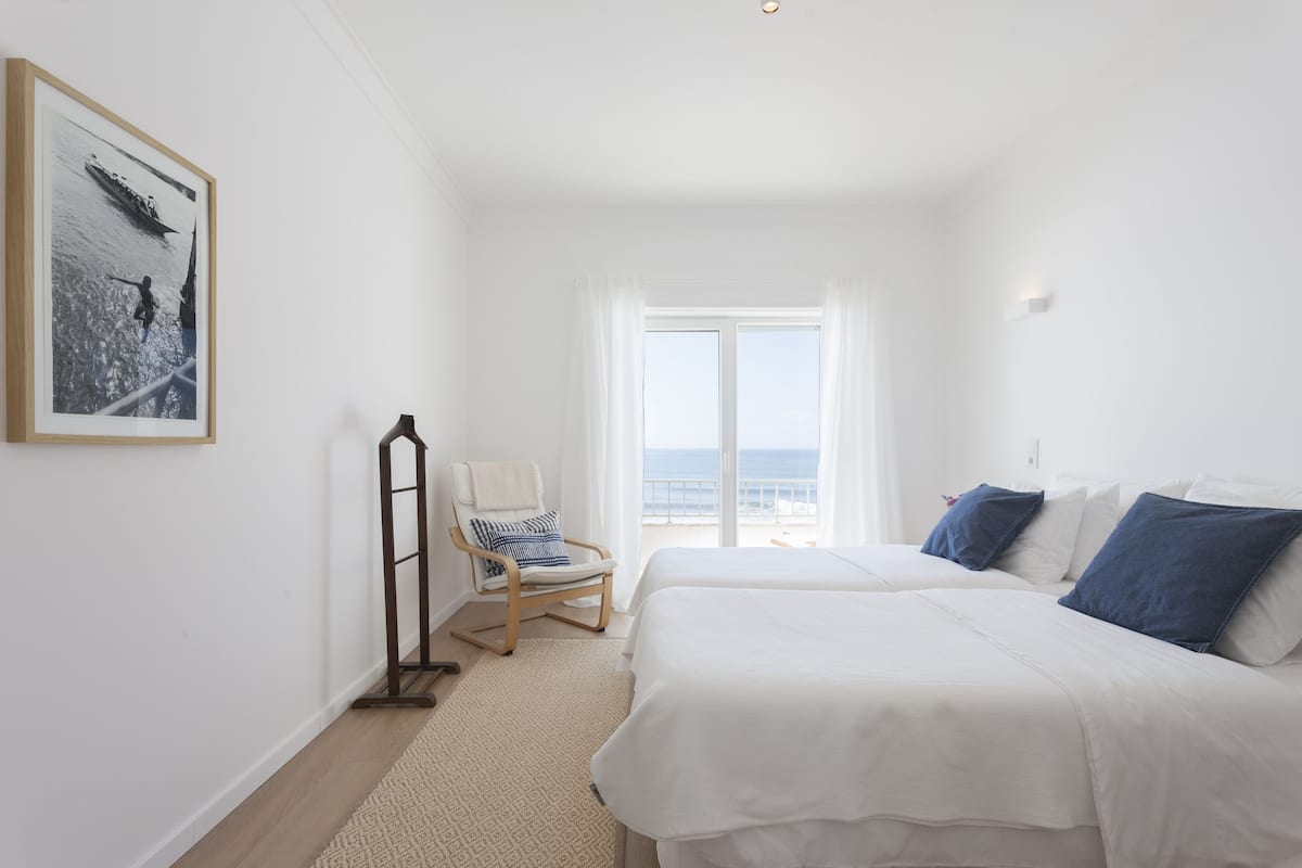 Een slaapkamer met een groot bed, een stoel, een lijst met een afbeelding en uitzicht op de zee door een raam.