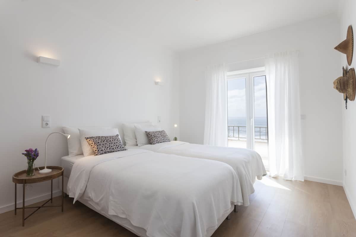 Een slaapkamer met twee bedden, een klein nachtkastje met een lamp, en uitzicht op zee door een groot raam.