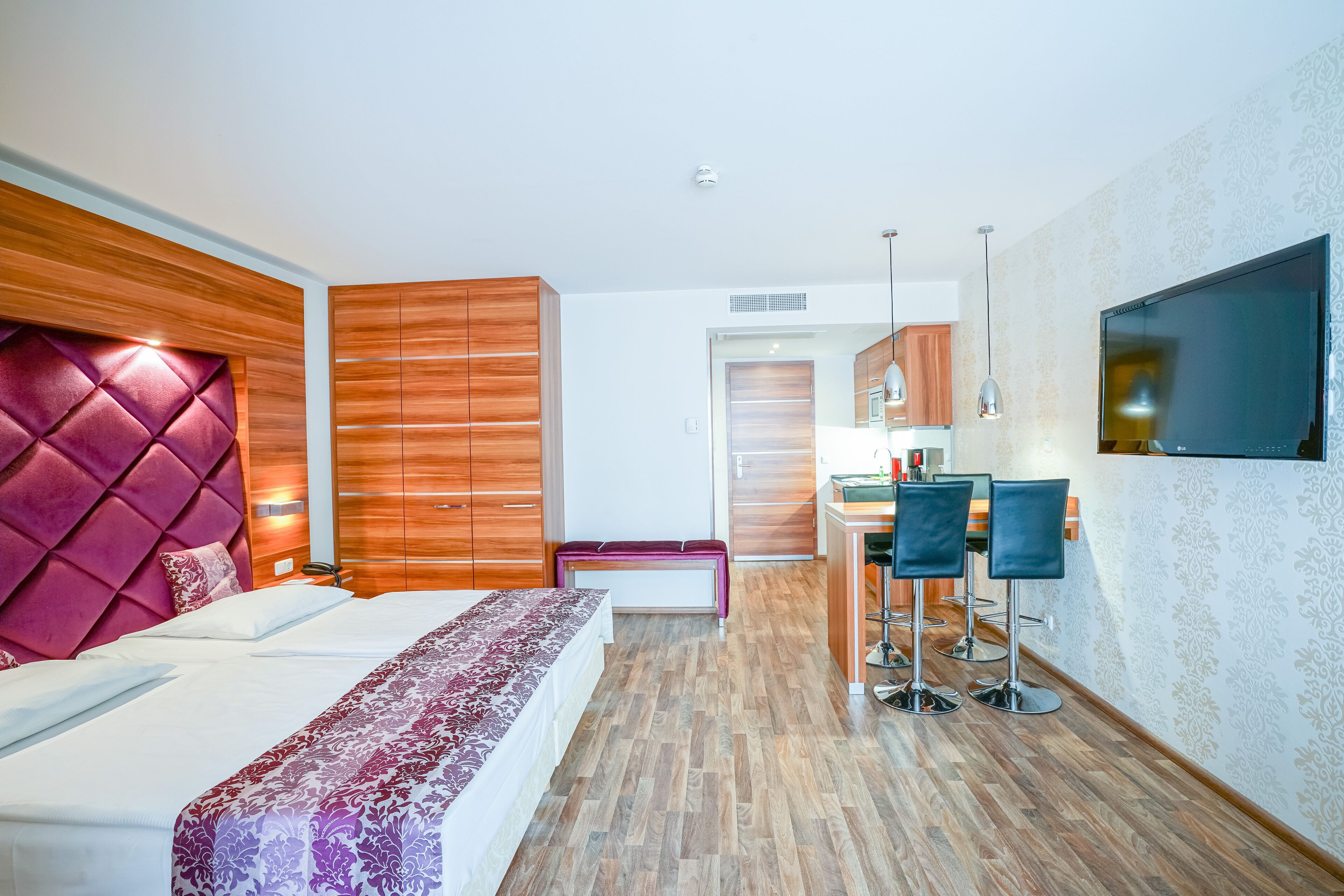 A modern hotel room with a large bed, wooden headboard, a bench, a desk with chairs, and a TV mounted on the wall.