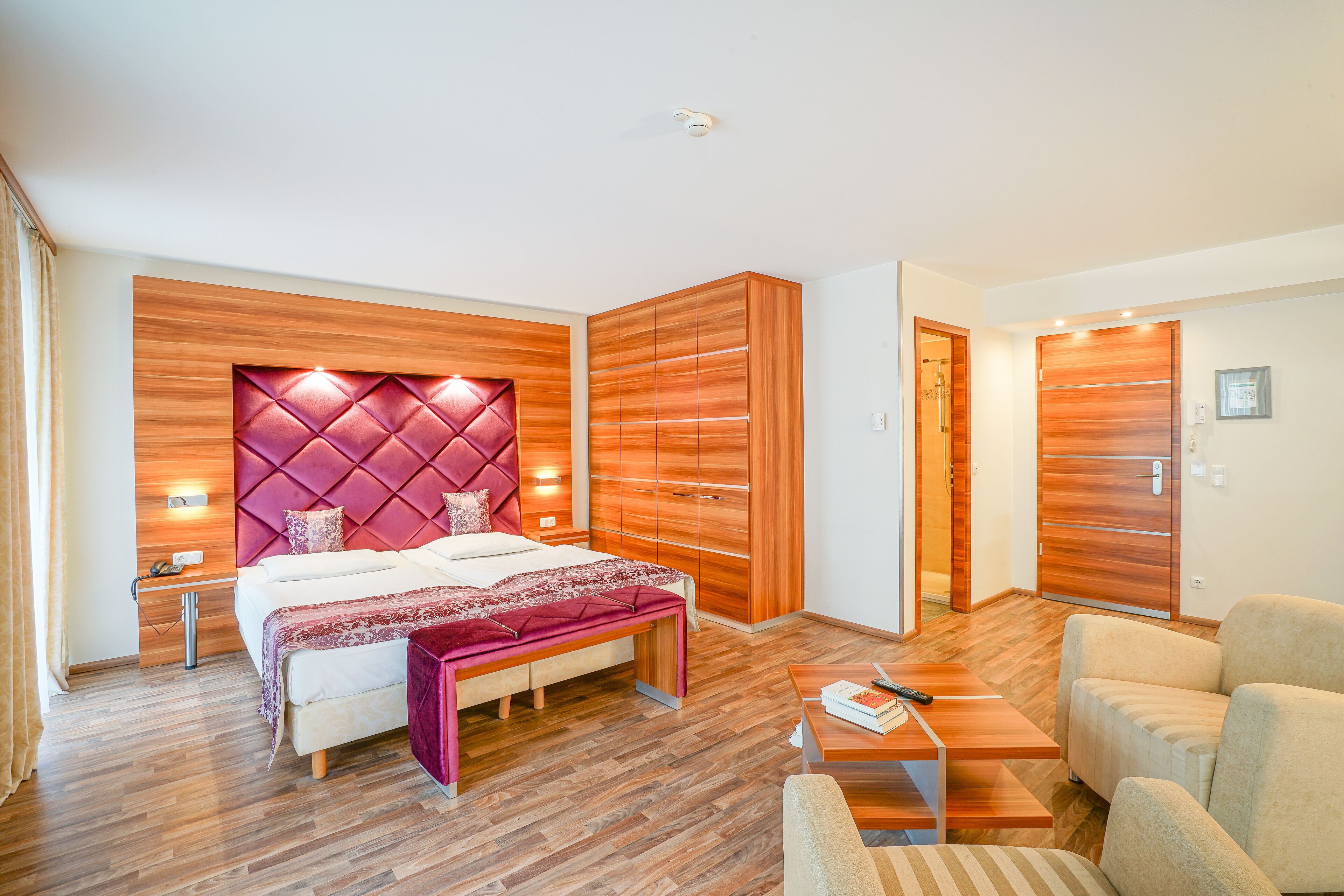 A modern hotel room with a large bed, wooden paneling, a bench, a desk, and a chair.