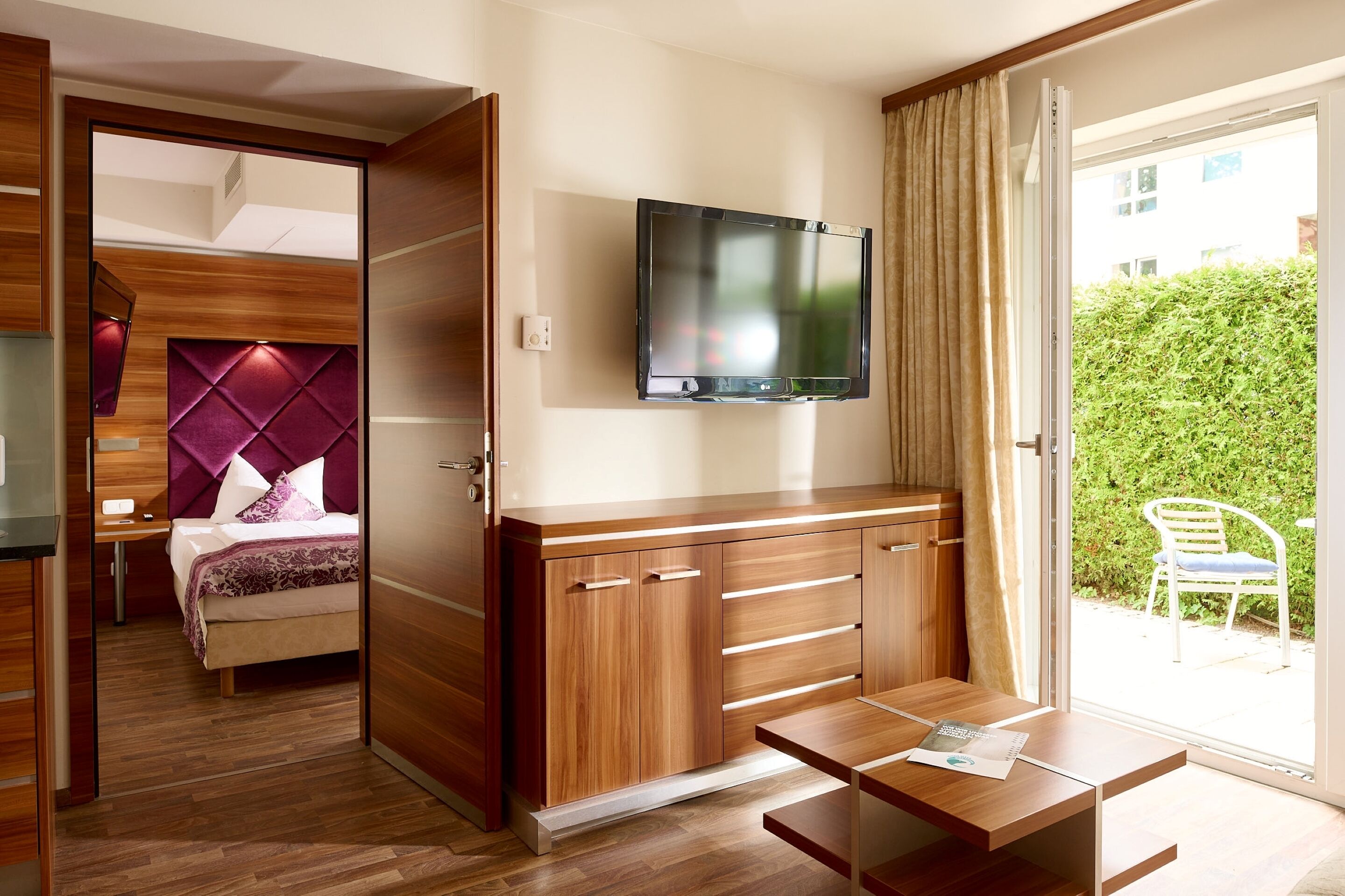 A modern hotel room with a wooden wardrobe, a bed with a purple headboard, a TV mounted on the wall, and a sliding glass door leading to a balcony with a chair.