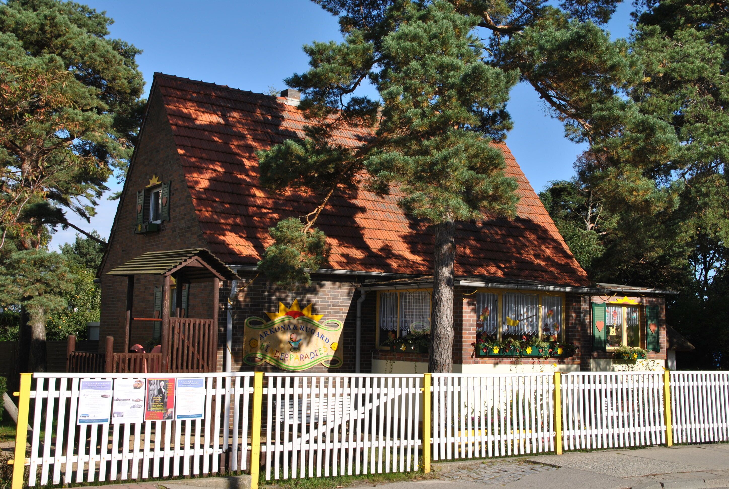 A brick building with a red roof, a white picket fence, and a sign that reads "ARKONA & RUGARD DER PARADISE".