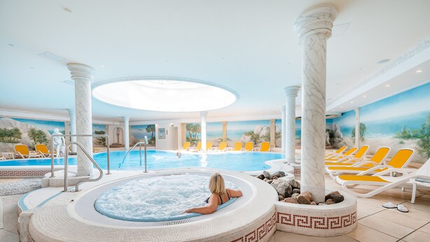 A large indoor swimming pool with a hot tub, surrounded by lounge chairs and columns. - Strandresidenzen Binz (Binz)