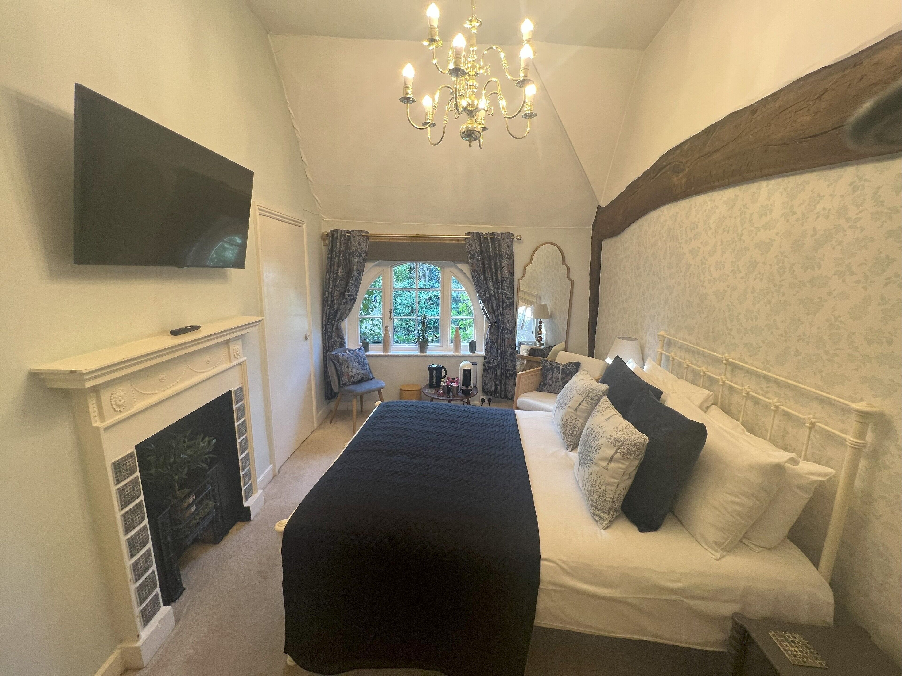 A bedroom with a bed, a television, a chandelier, a fireplace, a window with curtains, and a chair.