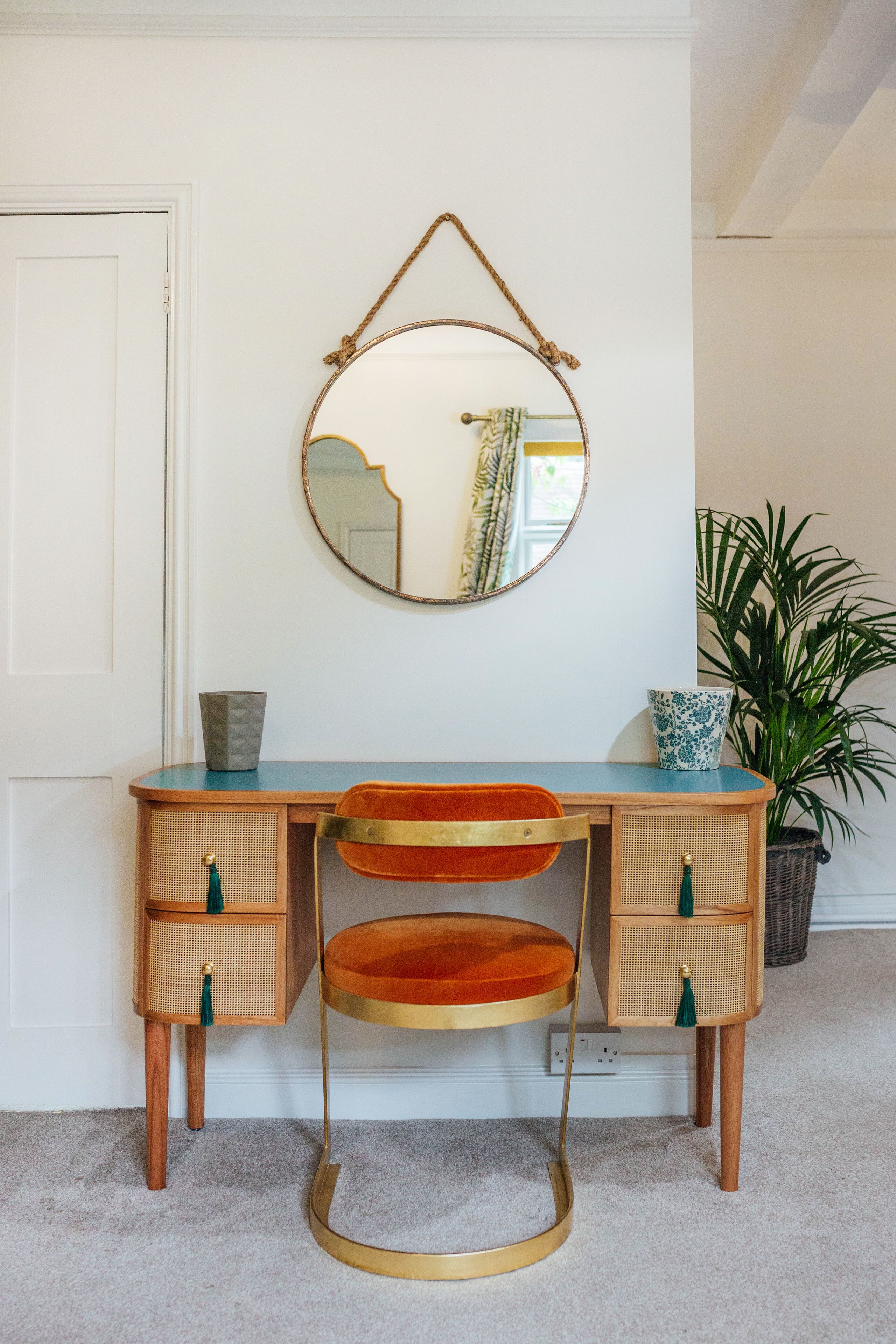 A vintage-style desk with a mirror above it, a plant to the side, and a small cabinet with drawers.