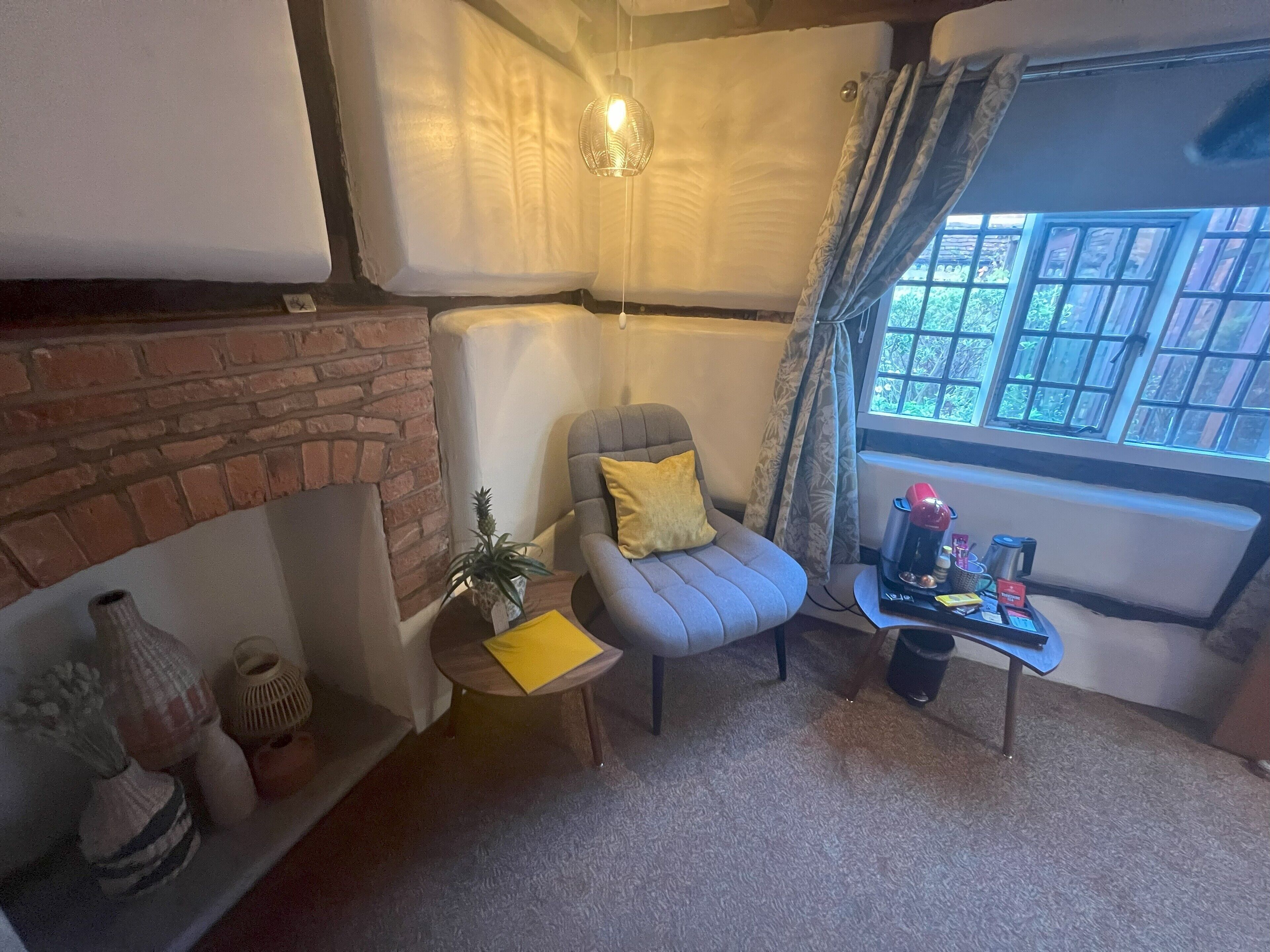 A cozy living room with a brick wall, a grey armchair with a yellow pillow, a small wooden table with a coffee maker, and a window with curtains.