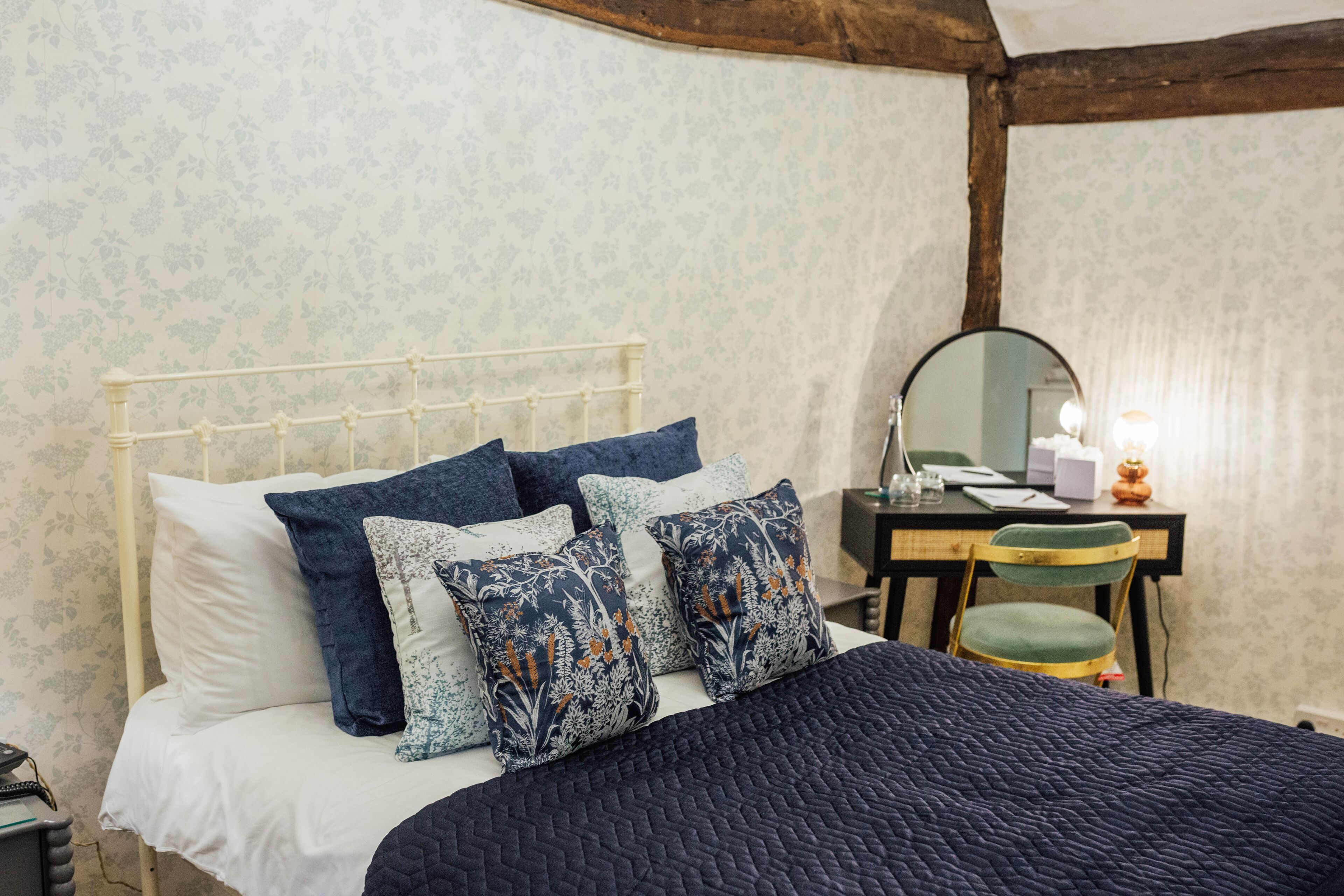 Una cama con cabecera blanca, almohadas azules con estampado floral y colcha azul oscuro. Hay un escritorio de madera con un espejo, una silla y una lámpara.
