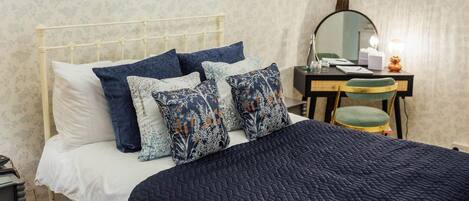 Una cama con cabecera blanca, almohadas azules con estampado floral y colcha azul oscuro. Hay un escritorio de madera con un espejo, una silla y una lámpara.