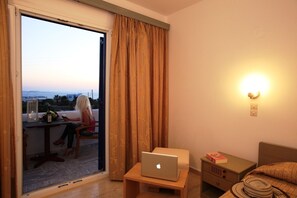 A room with a laptop on a small table, a nightstand with a book, and a view of the sea through a sliding door. - New Aeolos Hotel (Mykonos)