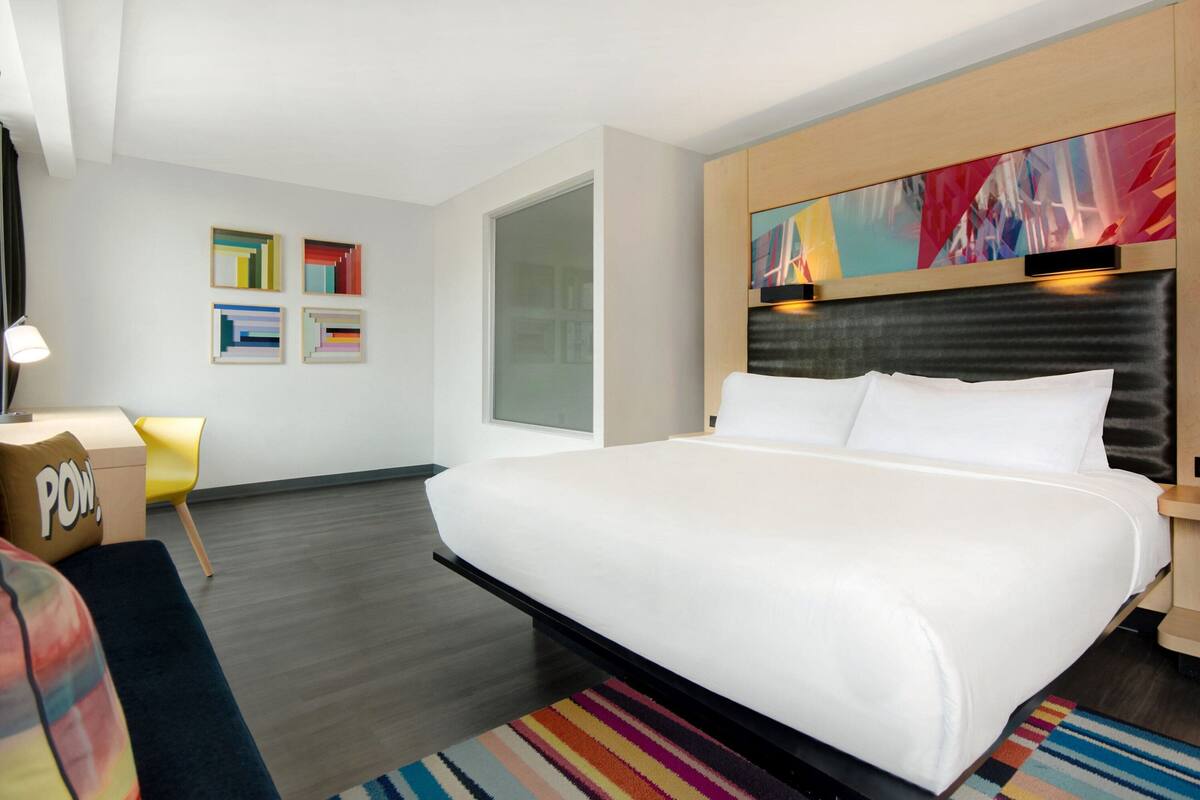 A modern hotel room with a large bed, a flat-screen TV, a desk, and a colorful abstract painting on the wall.
