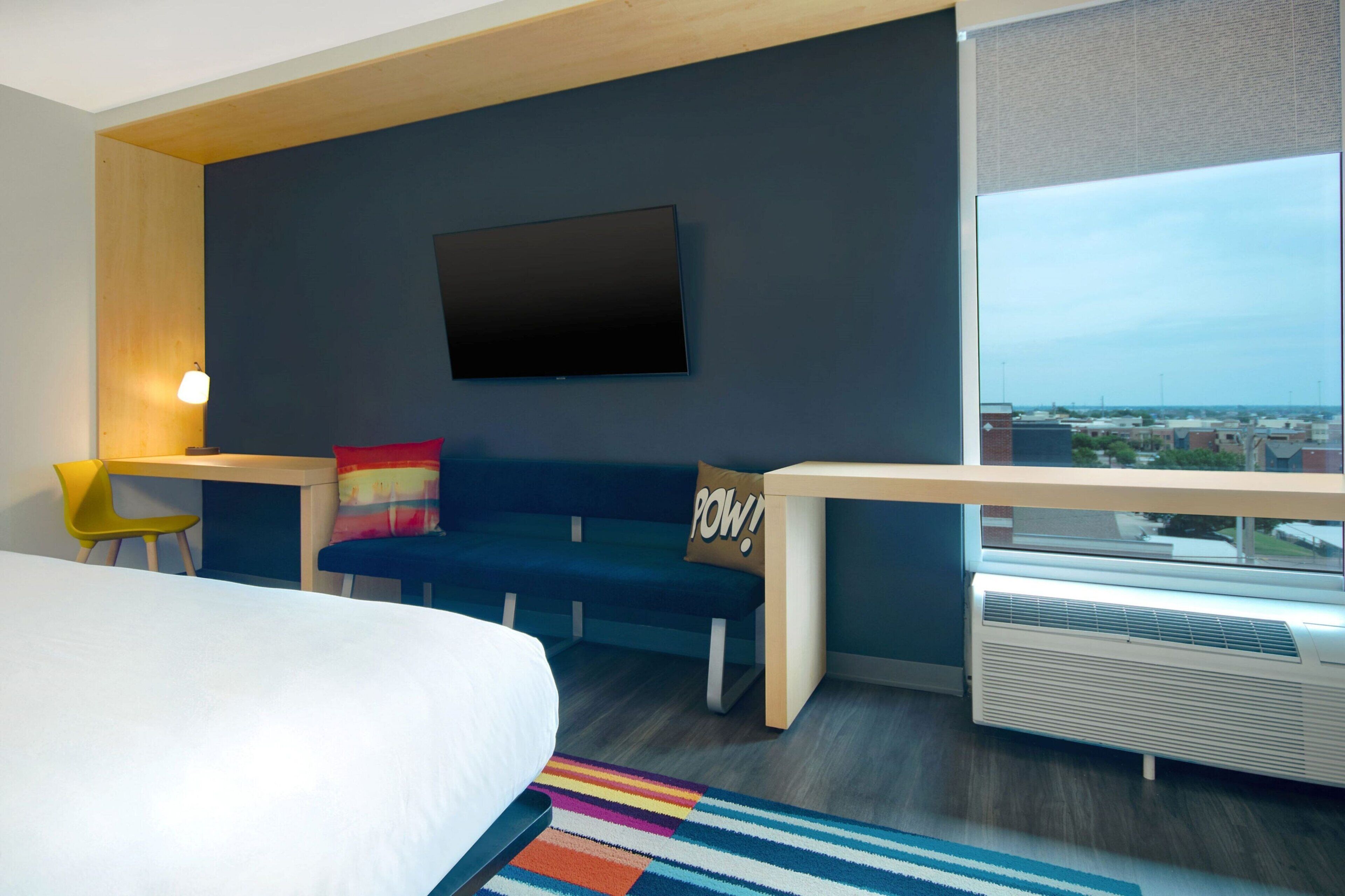 Una habitación de hotel moderna con una cama, un sofá, un televisor instalado en la pared, un ventanal con vistas a la ciudad y una mesita de noche con una lámpara.