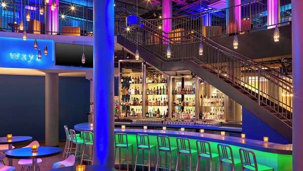 A bar area with multiple seating options, a staircase, and a variety of bottles on shelves. - Aloft by Marriott Oklahoma City Downtown - Bricktown (Oklahoma City)