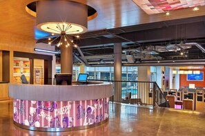 A modern hotel lobby with a central reception desk, a staircase, and a pool area visible through large windows. - Aloft by Marriott Oklahoma City Downtown - Bricktown (Oklahoma City)
