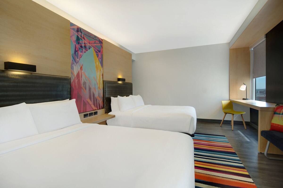 A hotel room with two beds, a desk, a chair, a colorful rug, and a large abstract painting on the wall.