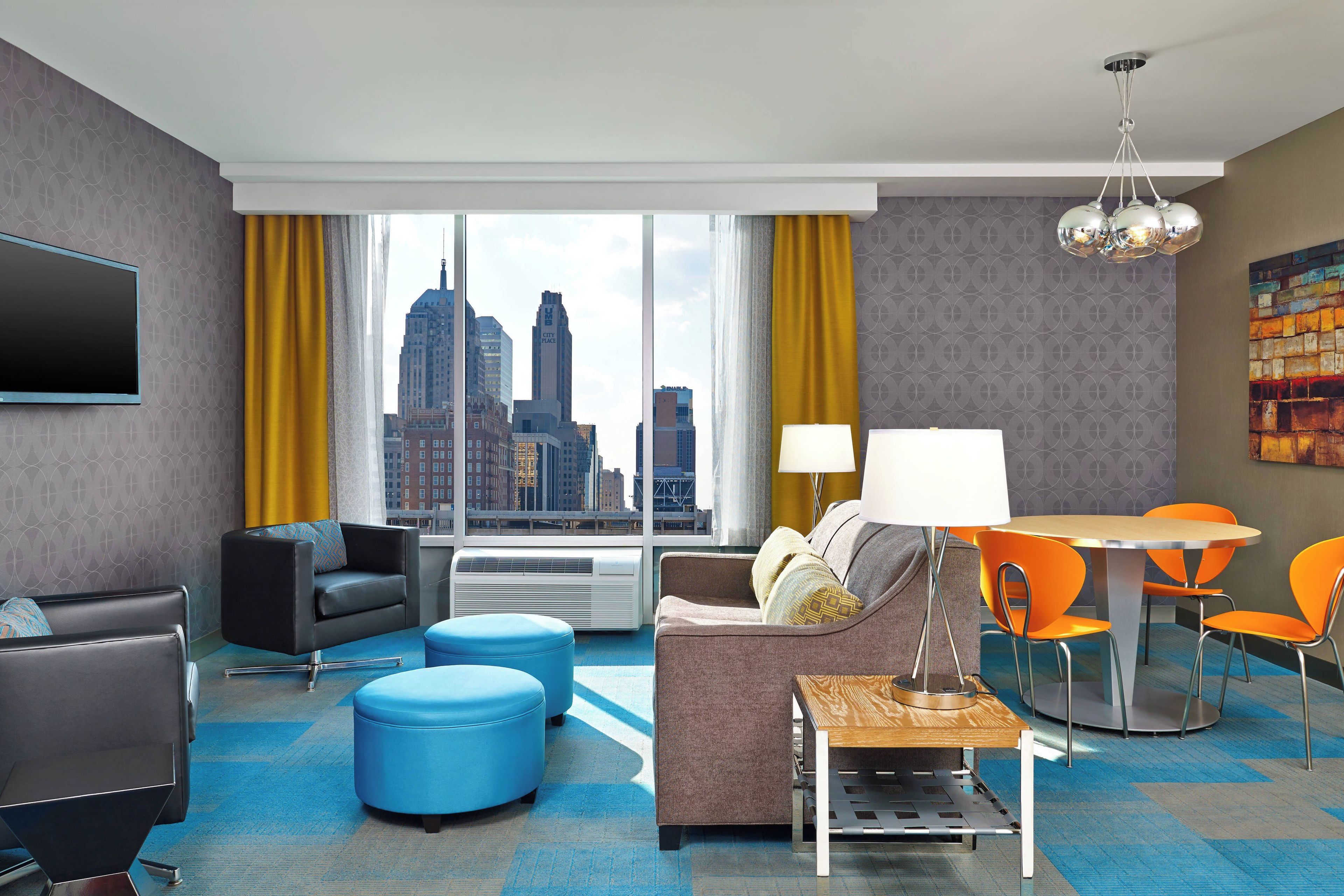 A hotel room with a city view, a flat-screen TV, a sofa, ottomans, a dining table with chairs, and a lamp.