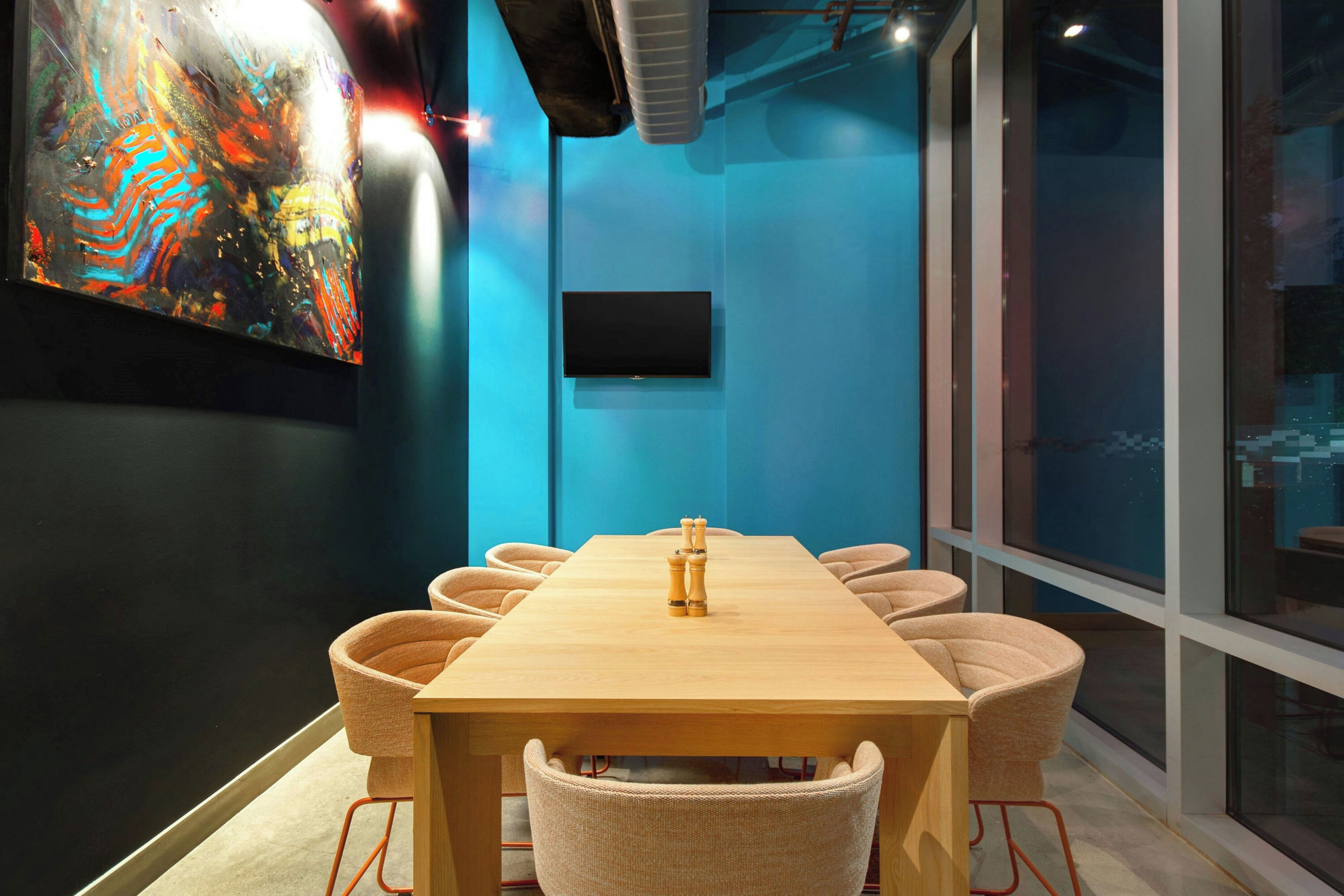 A conference room with a long table, six chairs, a flat-screen TV mounted on the wall, and a colorful abstract painting on the left.