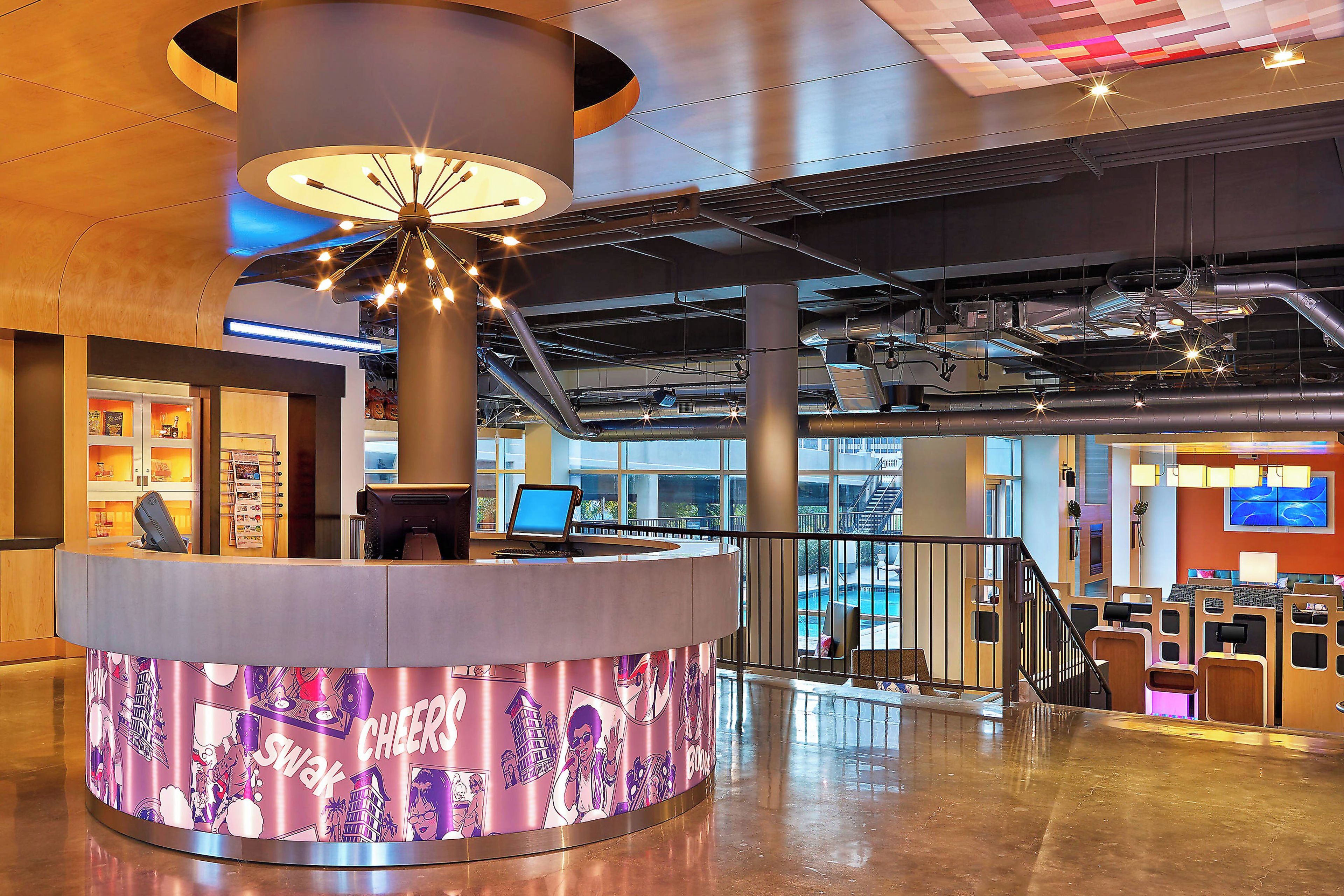 A modern lobby with a central reception desk, a staircase, and a pool area visible through the windows.