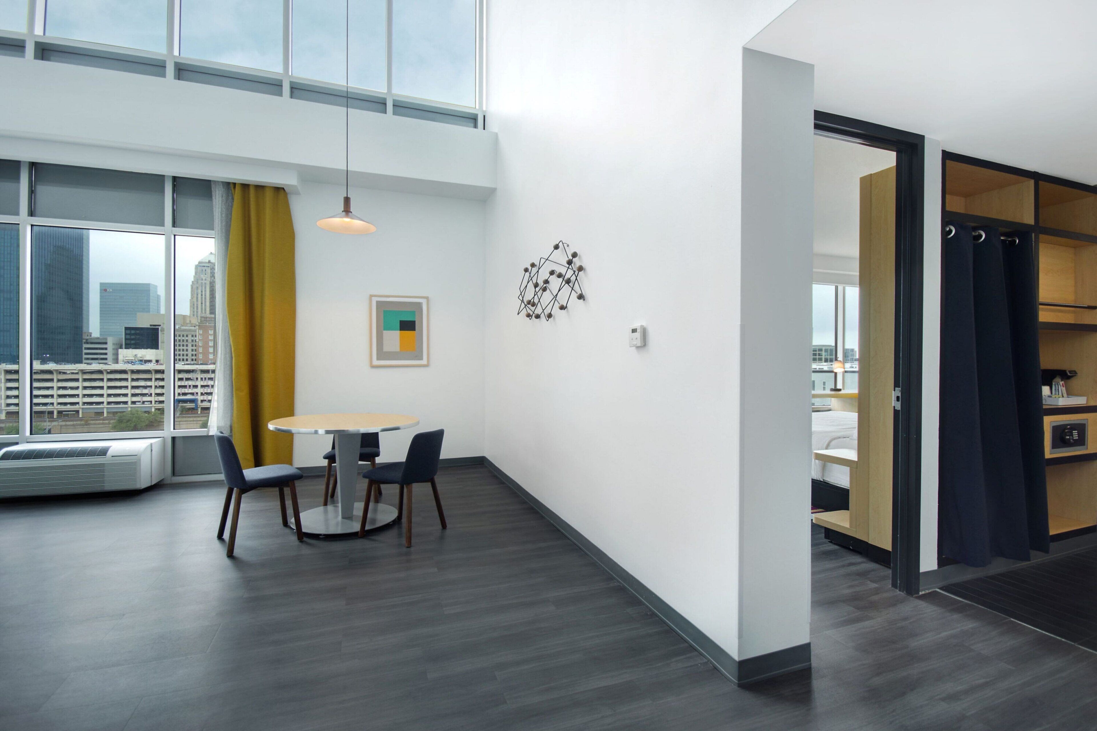 A modern hotel room with a large window, a dining area with a table and chairs, and a built-in wardrobe.