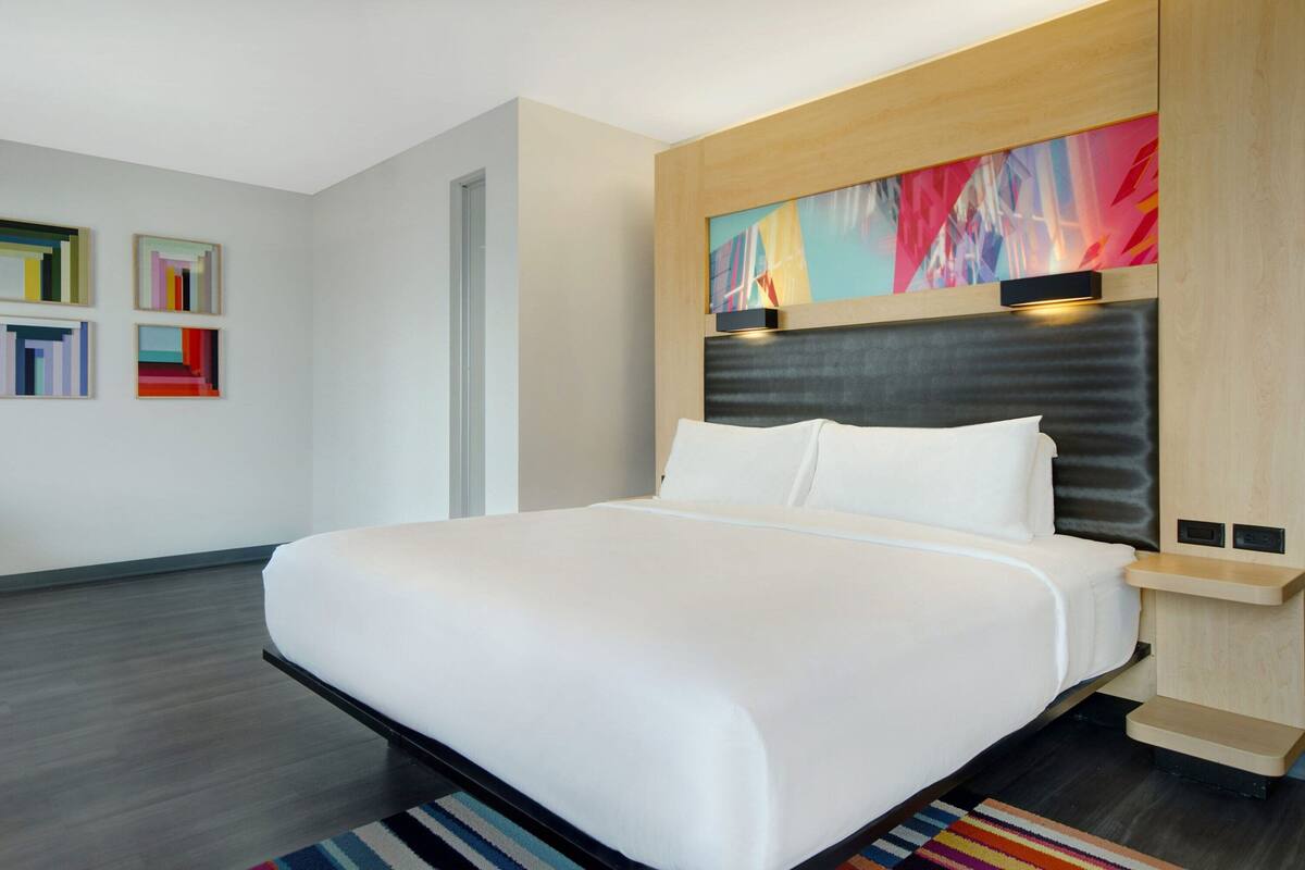 A modern hotel room with a large bed, a headboard with built-in lighting, a colorful abstract painting above the bed, and a small bedside table.
