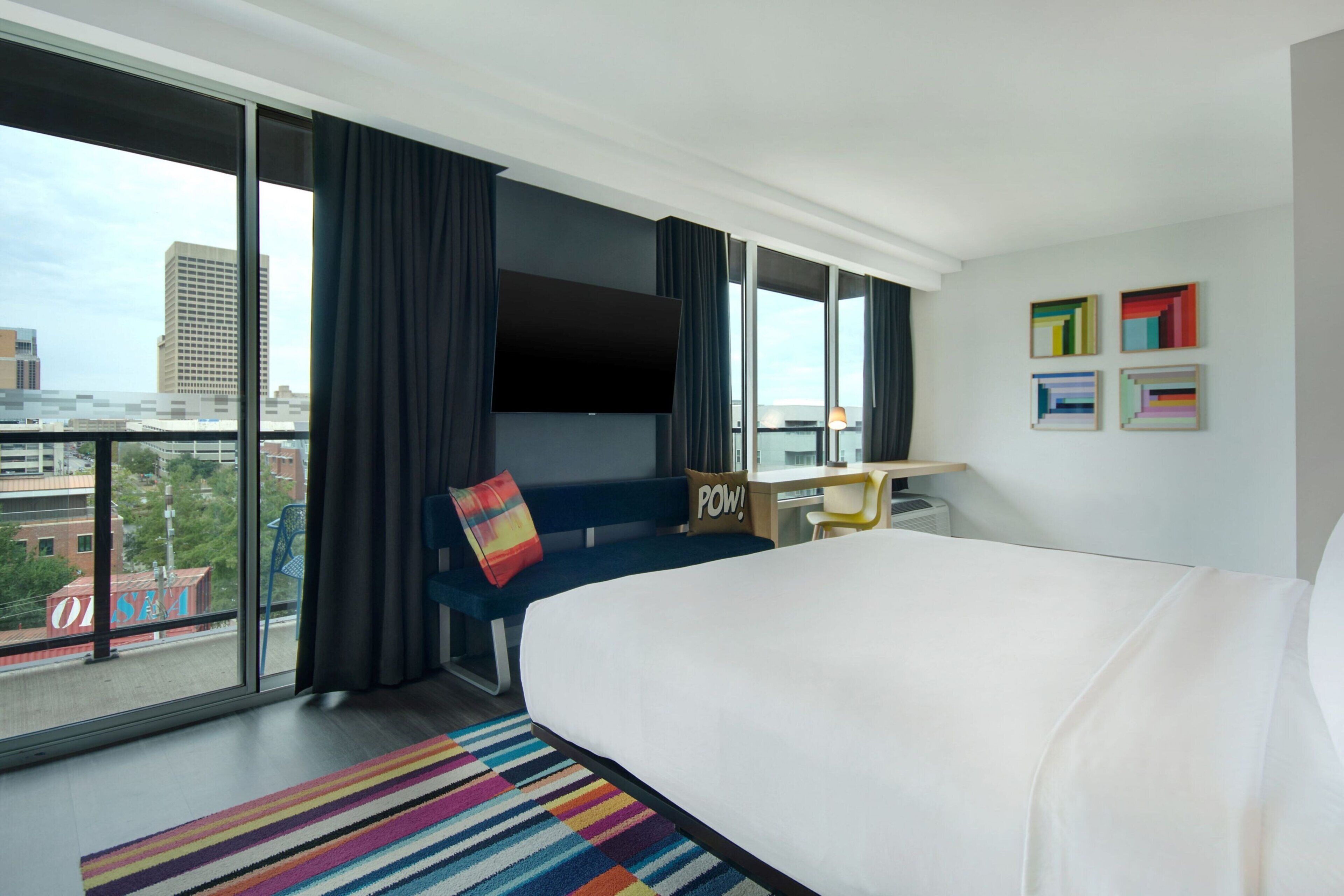 Habitación de hotel con una cama grande, un escritorio, una silla, un tapete de rayas coloridas, un ventanal con cortinas y vistas a la ciudad.