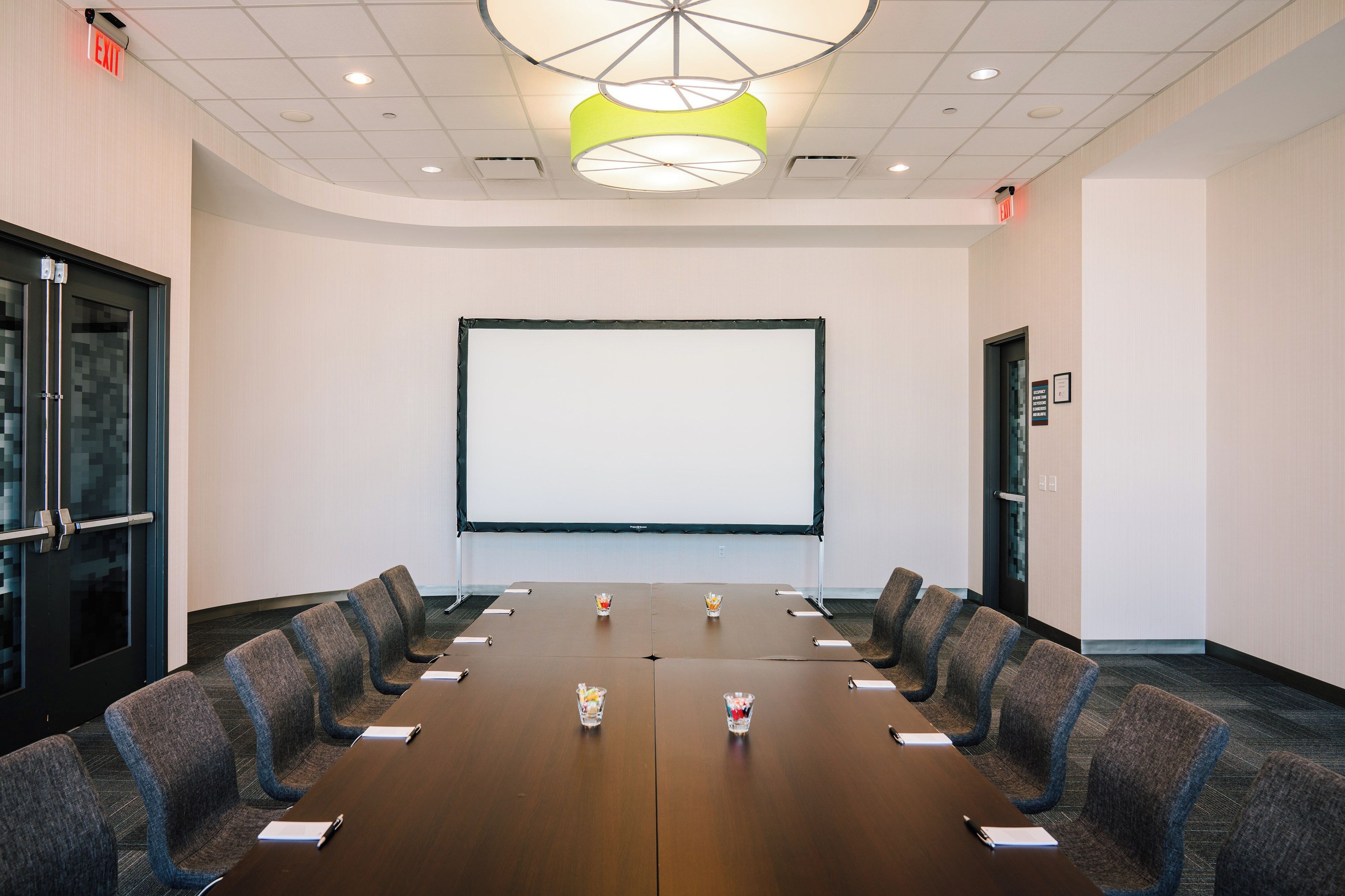 A conference room with a long table, chairs, a projector screen, and a ceiling light fixture.