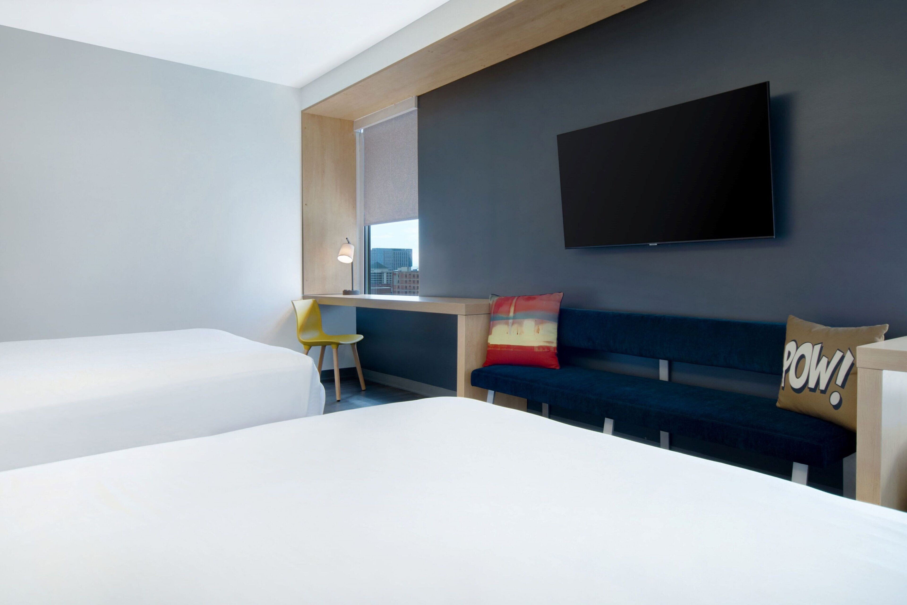 A modern hotel room with a large bed, a desk with a chair, a flat-screen TV mounted on the wall, and a sofa with a 'POW!' pillow.